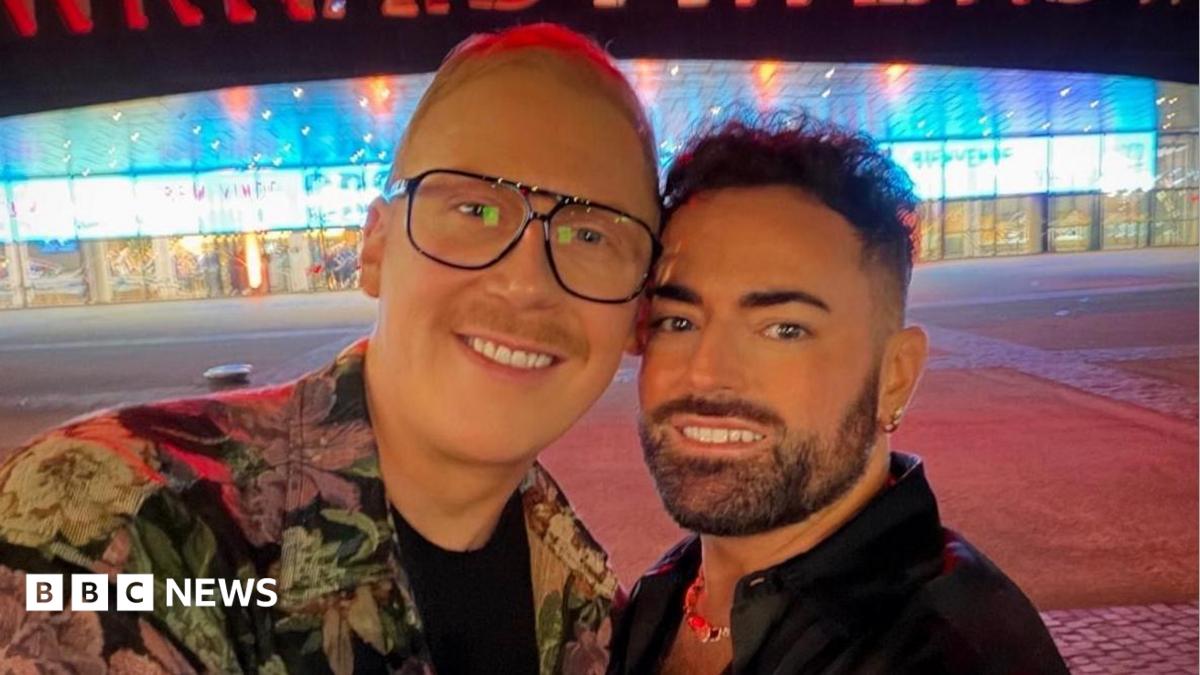 Karl, who has dark hair and a short beard, smiles next to his partner Craig who has blonde hair and is wearing a floral shirt and glasses