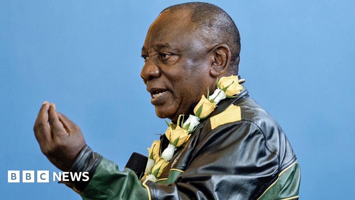 Cyril Ramaphosa, wearing a leather green, yellow and black leather jacket and with a yellow and white rose garland around his neck, gestures with his hand as he talks.