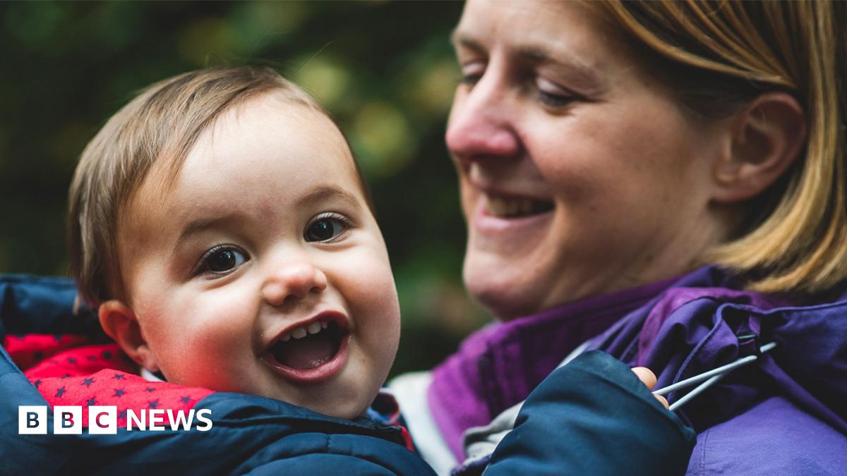 James, pictured as a young child, is held by his mother Laura in an outdoor setting, with greenery in the background.