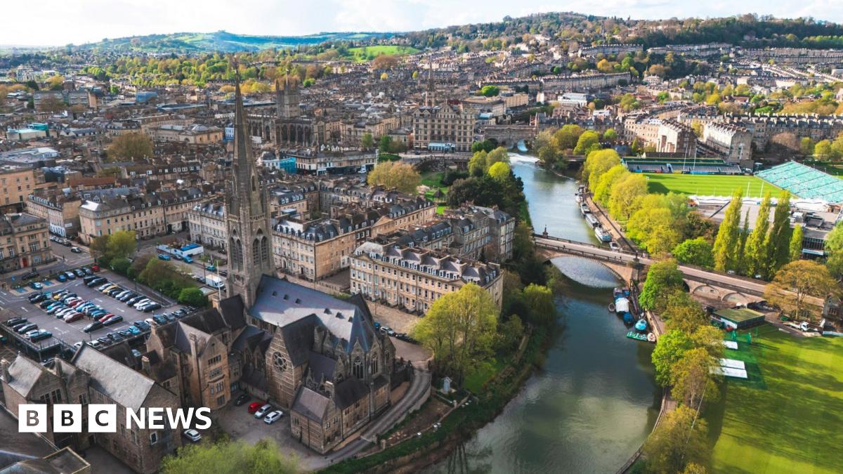 An aerial view of Bath city centre taken by a drone. Landmarks such as the River Avon and Bath Rugby's Rec stadium are visible and it is a clear, sunny day with a range of hills in the background