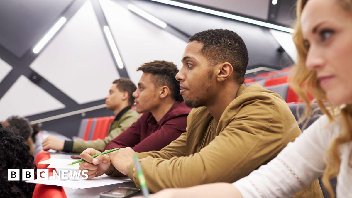 A stock photo shows four students sat in a row of a lecture hall, as taken from the side. Three are men and there is a young woman in the foreground. They have papers in front of them and pens in their hands, and are looking intently to the left, towar...