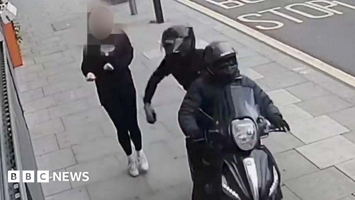 A CCTV image showing a black moped being driven along a pavement by two people wearing all black, snatching a mobile phone from a pedestrian walking along the pavement. One of the people on the moped can be seen holding the phone in their hand moments...
