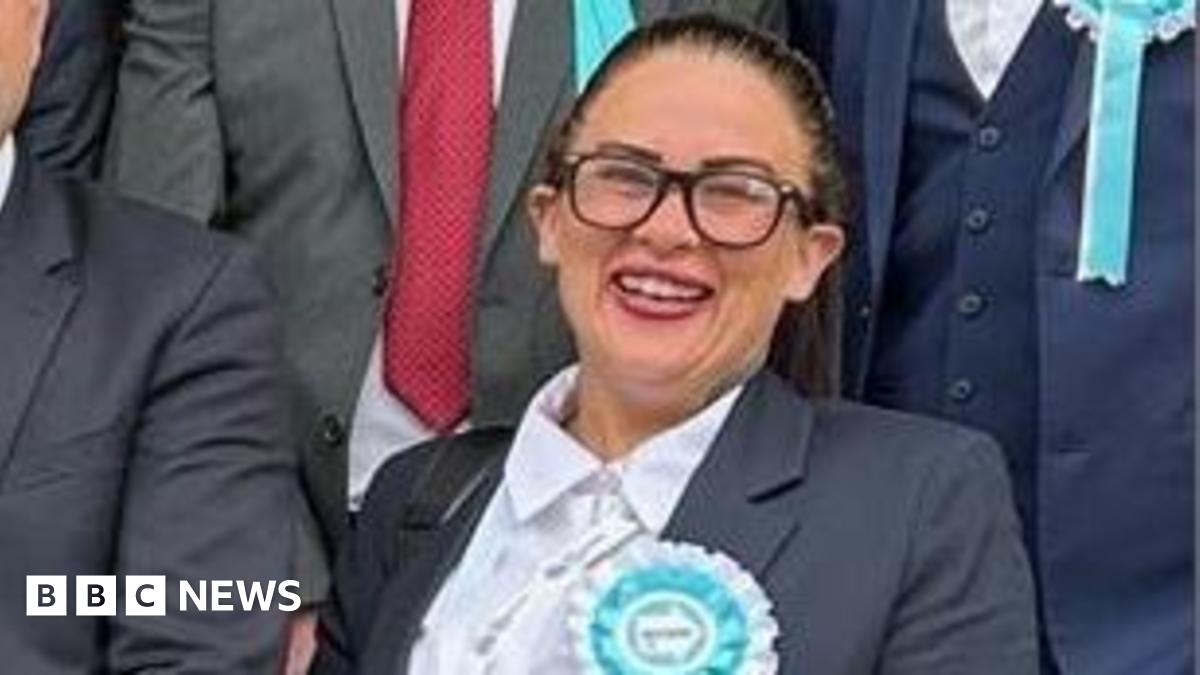 Kathryn Shaw stands next to council leader, Mark Arnull. They both wear suits and Shaw has a pale blue rosette indicating Reform UK on her jacket lapel. Other members of the Reform Party are stood behind them but you cannot see their faces.
