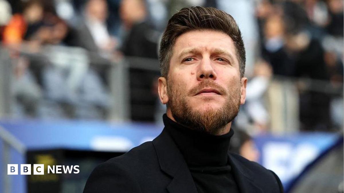 Photo: bbc.co.uk A man wearing a black polo neck and blazer with a quiffed hairstyle and a beard. He is standing in front of a stand full of