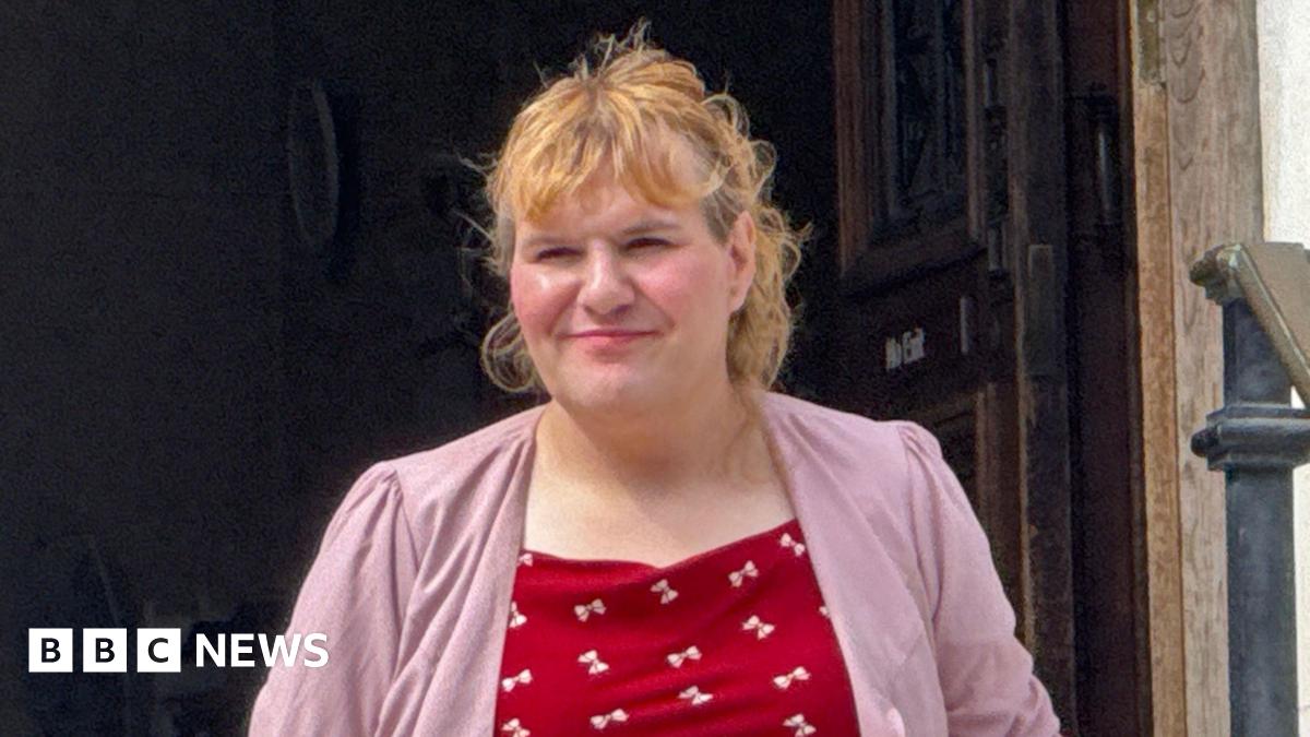 Katie Wallis walking out of court building wearing red top with white bows and pink blazer over it. She has long curly blonde hair up in a pony tail.