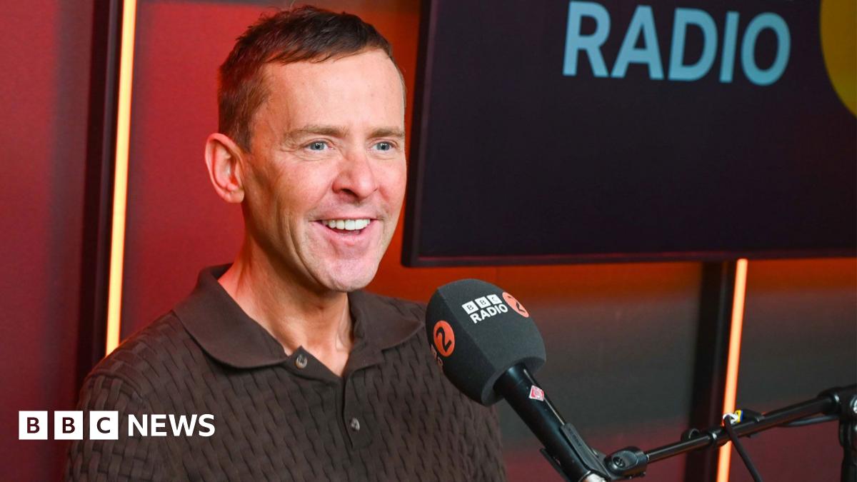 Scott Mills in a Radio 2 studio, he is wearing a brown polo top