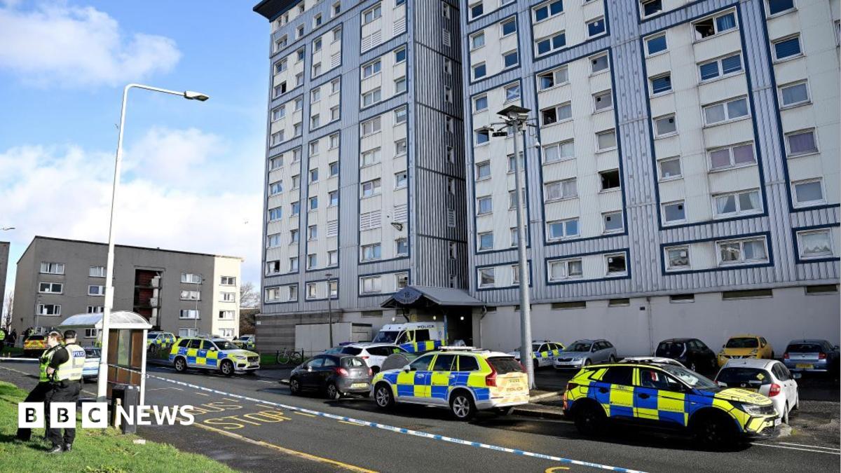 A block of flats clad in blue and white. Police cars line the residential streets and there is police tape blocking off the area, tied to a lamppost. Two police officers stand on the pavement.