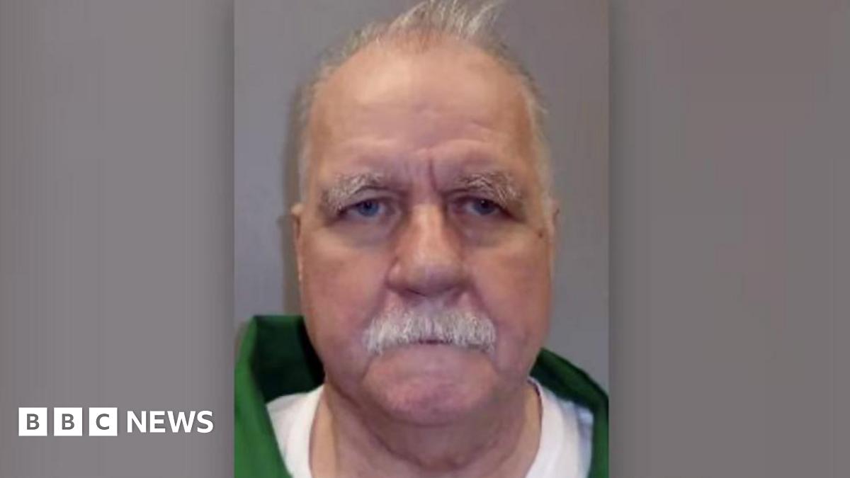 The prisoner is seen in a green jacket with a white shirt. He has a white mustache and white hair and there is a grey background