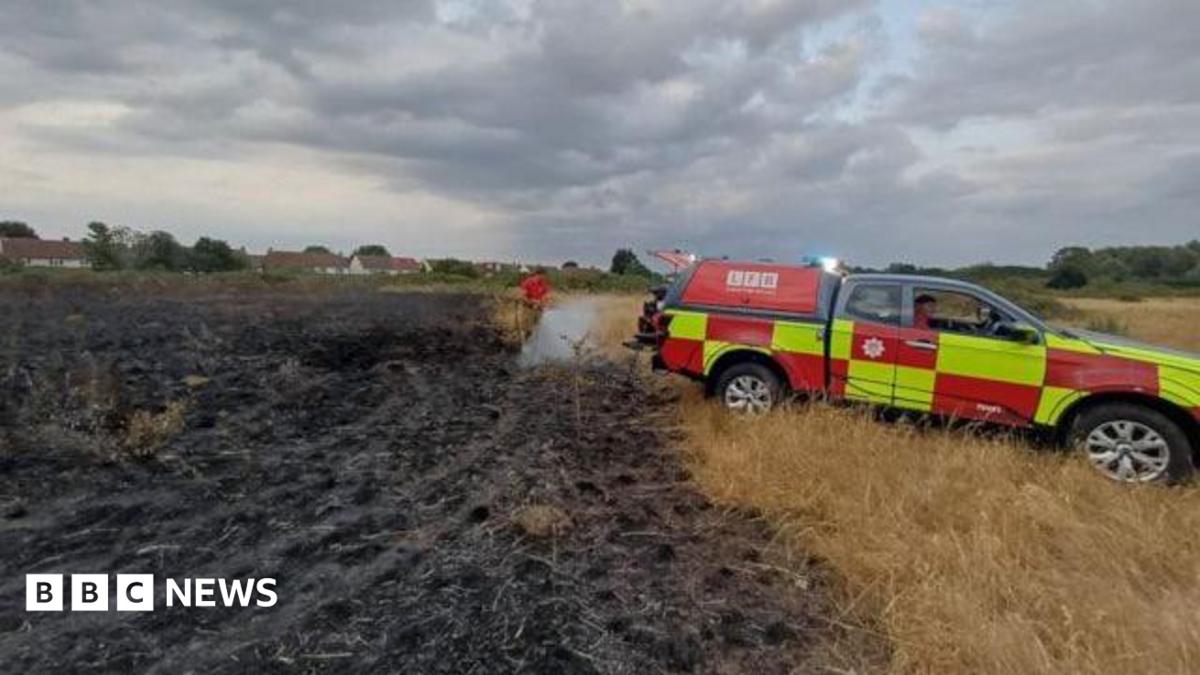 An all terrain fire brigade vehicle parked by a burnt area of grassland. A firefighter is hosing down undamaged grassland adjacent to the burnt area. 