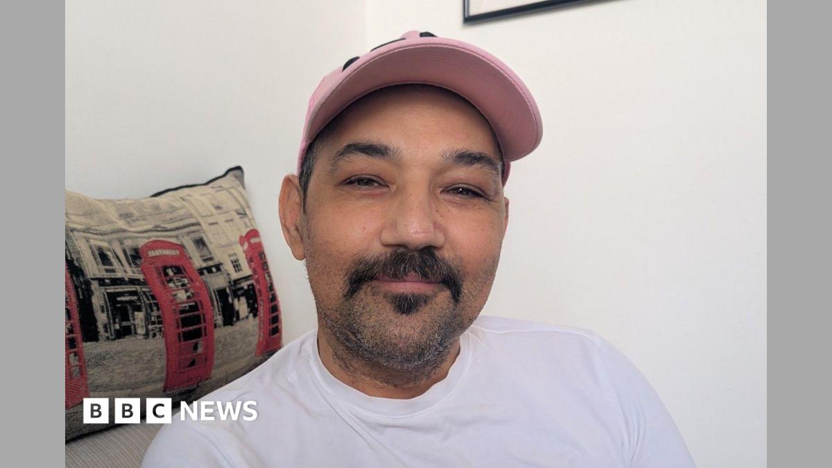 Photo: bbc.co.uk A young man wearing a white t-shirt and pink baseball hat seated indoors next to a decorative cushion featuring iconic red