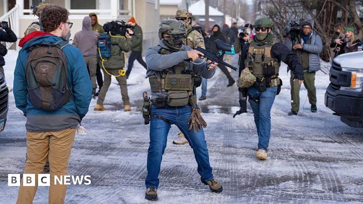 An officer with US Immigration and Customs Enforcement or ICE (right) and another federal officer holding a crowd control device (centre) stand at a Minneapolis intersection where protesters had gathered after the death of Renee Nicole Good. Bystanders...