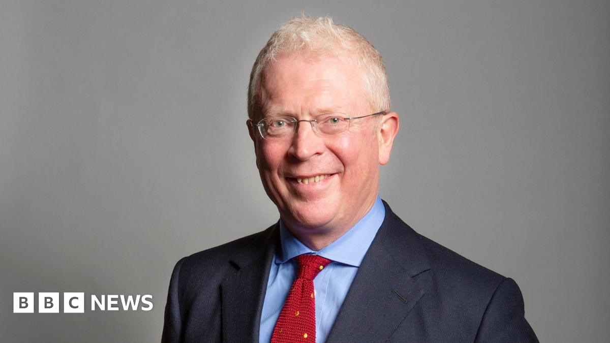 A bespectacled man in a navy suit, blue shirt and red tie.