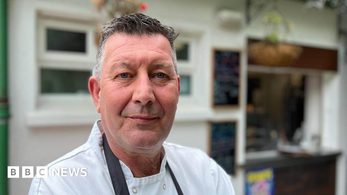 A man with grey hair, wearing a white chef shirt and a dark blue apron, is looking into the camera. He's standing in front of the cafe he owns, and there are two black boards on the white building wall behind him and a hatch for serving.