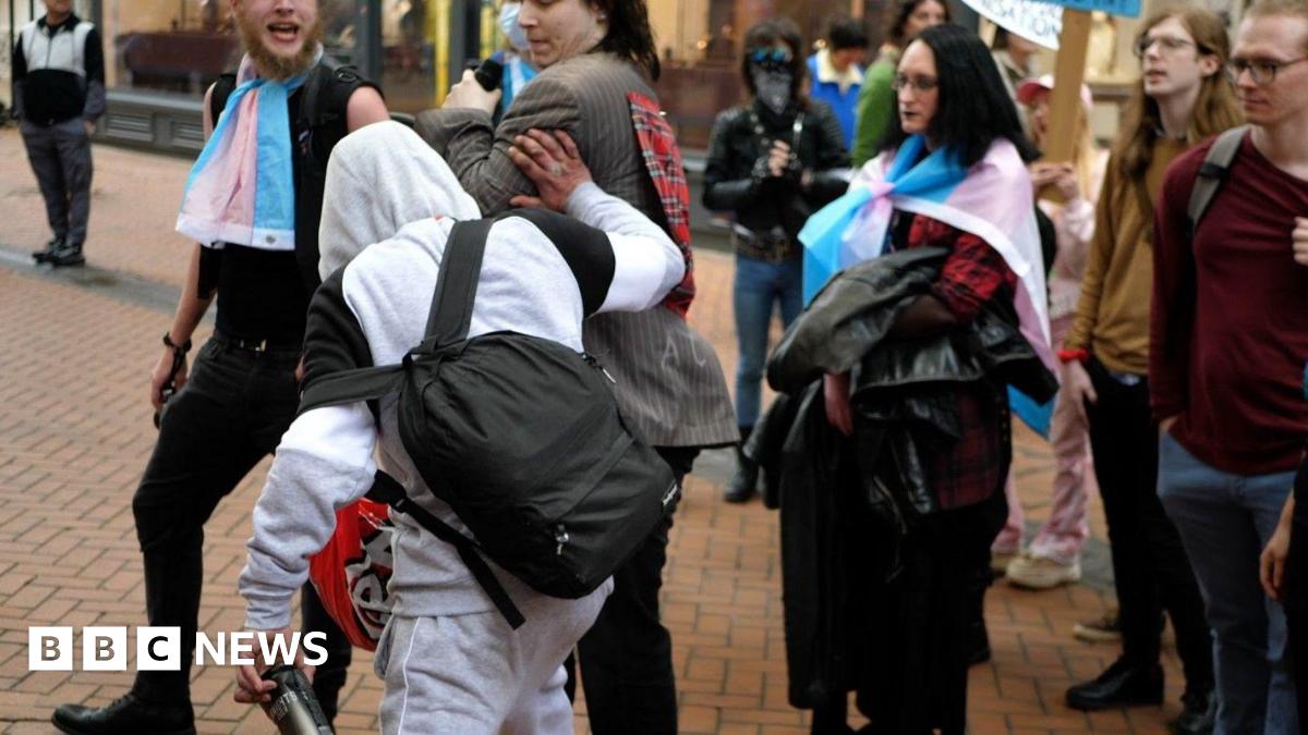 A man in a grey hoodie, wearing a black rucksack and carrying a canned drink, can be seen pushing someone wearing a suit jacket while a group of people who appear to be involved in a protest are stood nearby.