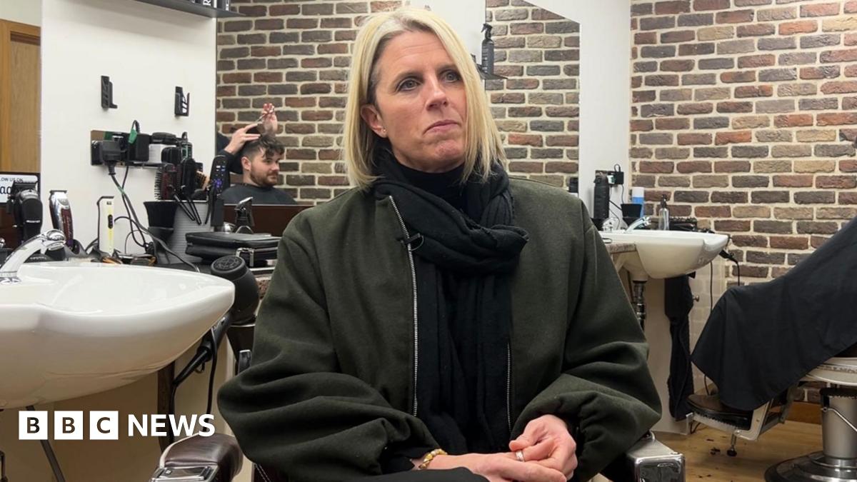 A close up shot of Lisa with long blond hair and a black jacket sat in a barber's with sinks around her and someone having their hair cut in the background