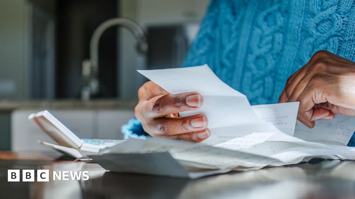 Close up shot of a person looking at bills and receipts on a table next to a calculator in their kitchen. They are wearing a blue jumper and the kitchen sink is in the background.