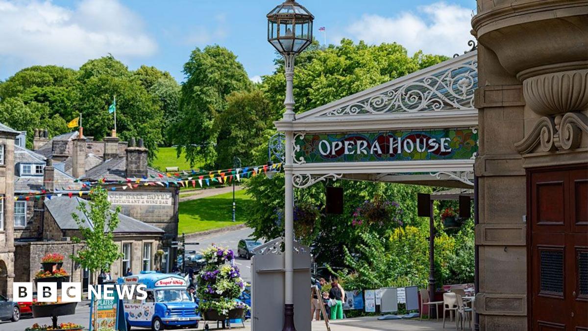 The exterior of a limestone-brick building with a veranda that says "opera house". Behind it, it's a sunny day, and there are trees full of leaves on a green hillside.