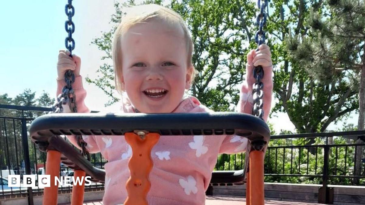 A little toddler on a swing. She has short wispy blonde hair and is beaming at the camera.
