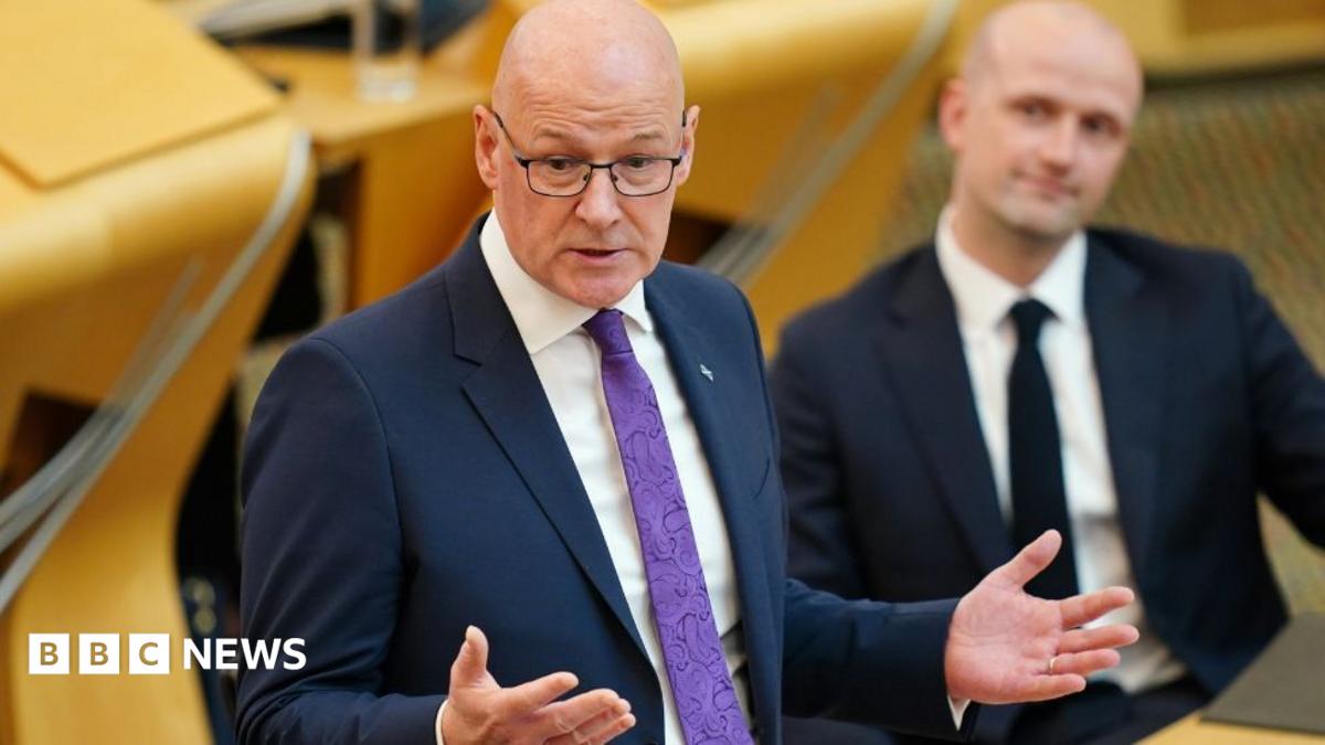 John Swinney, who is bald with glasses, speaks into a microphone while wearing a business suit 