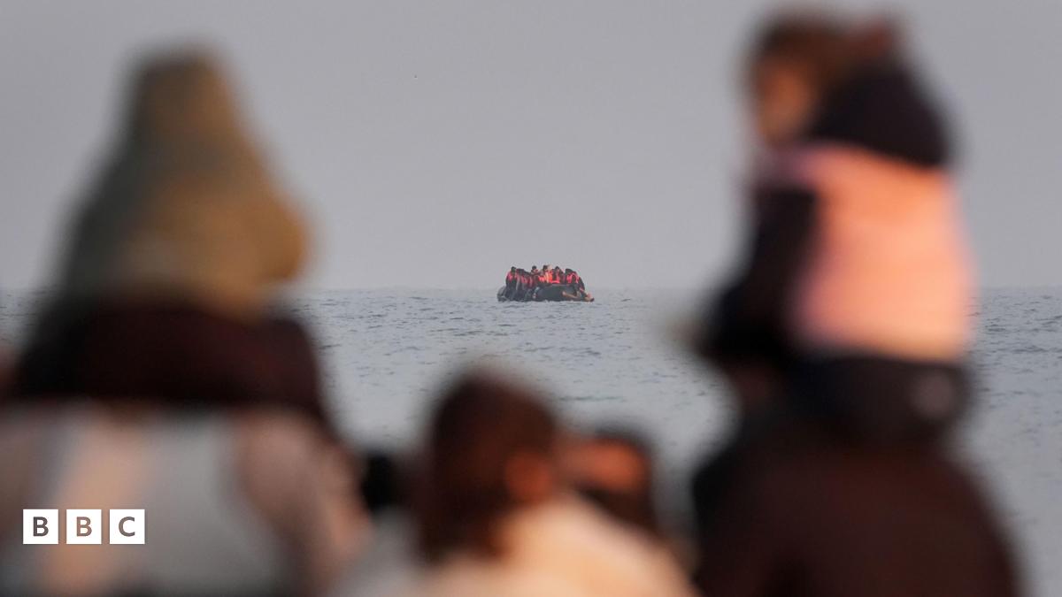 In the foreground are two children, carried on the shoulders of adults. They are pictured out of focus, looking out to sea. In the distance, a small boat full of people wearing life jackets is seen at sea.