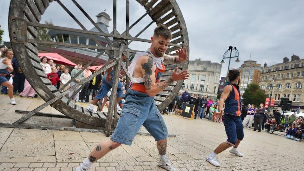 Two performers stand in front of a large wooden wheel with another inside it.  A crowd looks on.