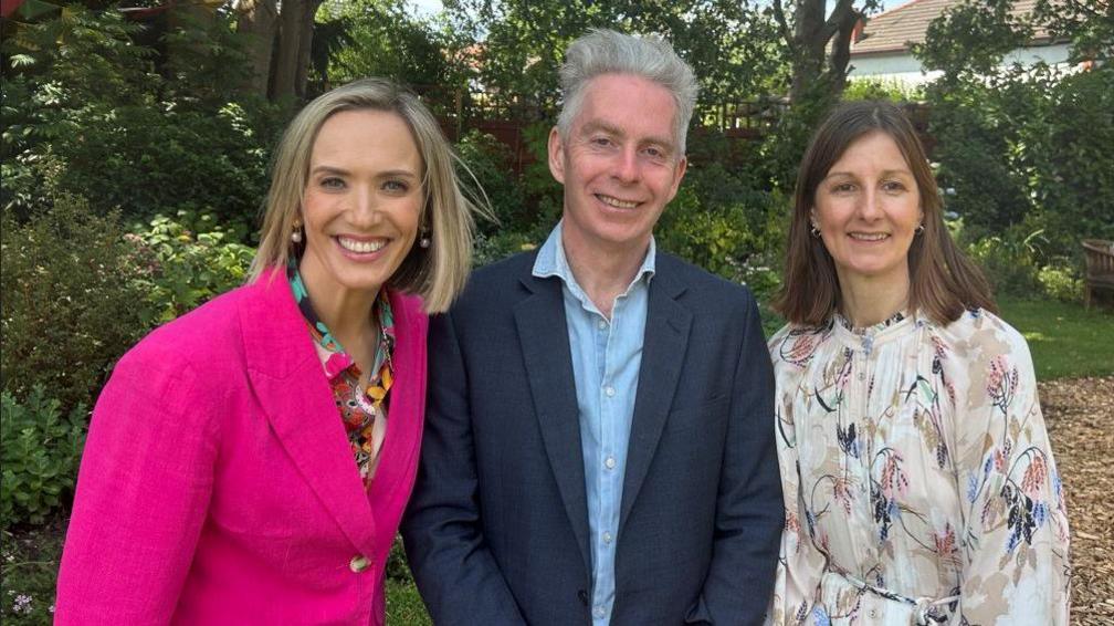 Kay Crewdson is standing next to John Sainsbury and Sheila Ryan in the gardens of the centre. Flowers, plants and trees are in the background. All three are starring directly at the camera and are smiling.