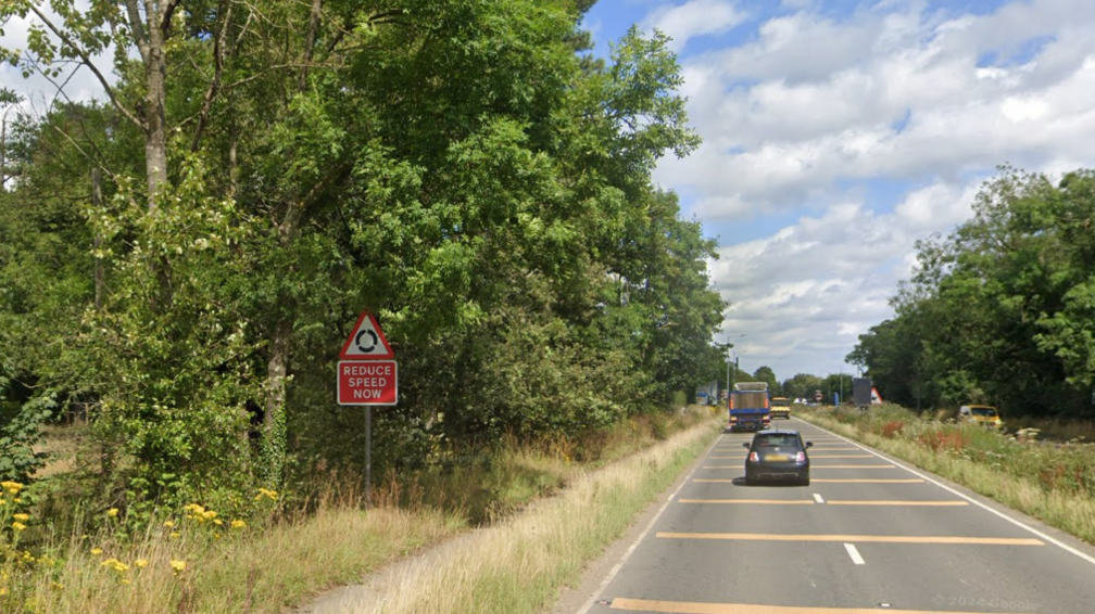 A rural dual carriageway pictured on a sunny day. A car and an HGV are visible. There is a sign reading "reduce speed now".