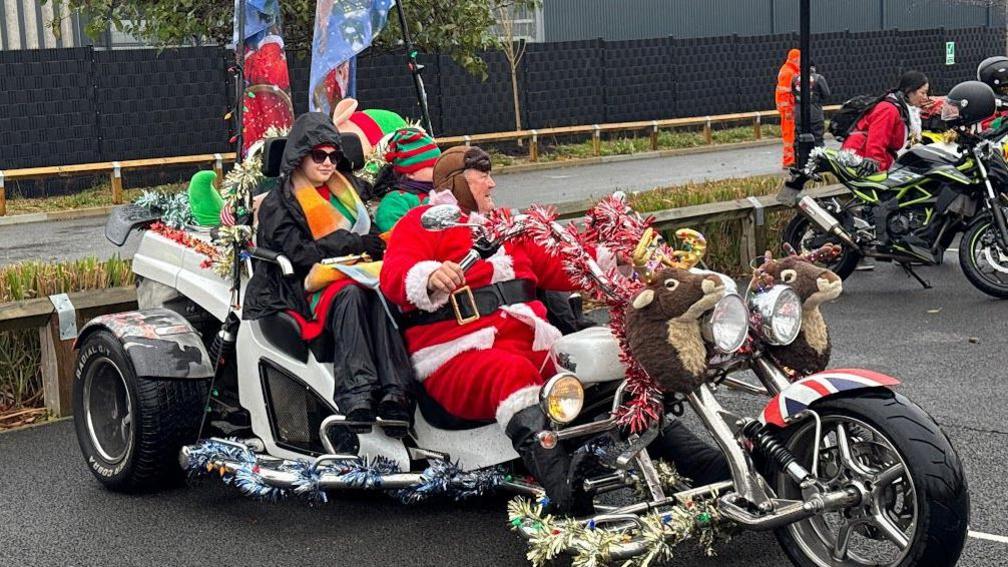 Three people sitting on a large trike covered in tinsel and soft toys. The rider is dressed as Santa.