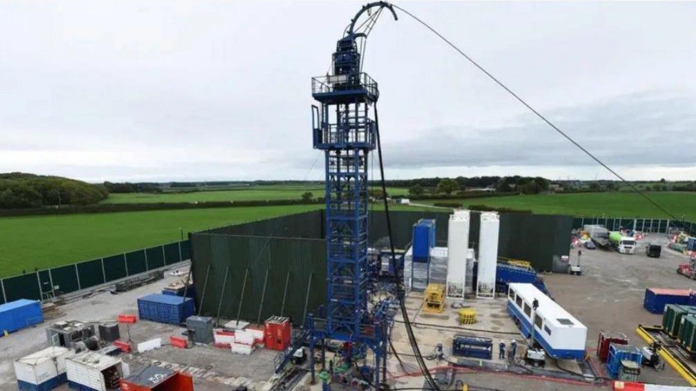 Fracking equipment and various storage units can be seen on the site, which is surrounded by green fencing.