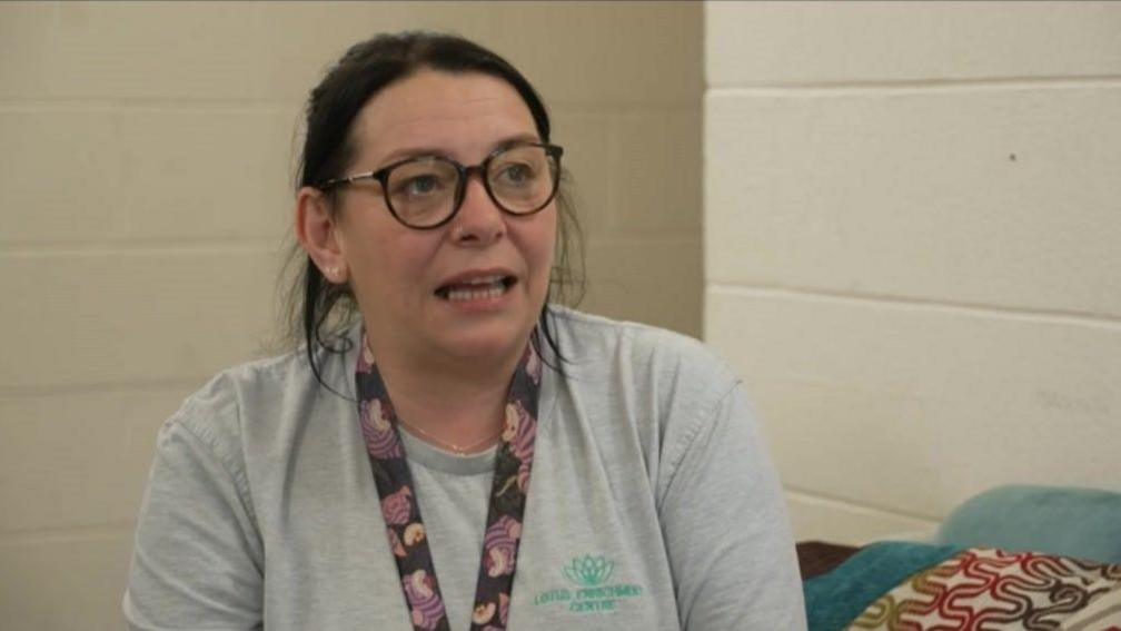 A woman with brown hair and glasses wearing a grey T-shirt with the Lotus Enrichment Centre logo on it.