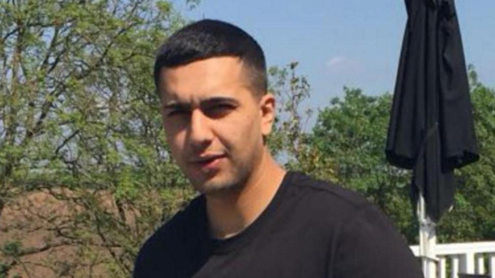 Image of Kamran Aman wearing a black t shirt and has short cropped hair. There are trees and a white railing behind him.