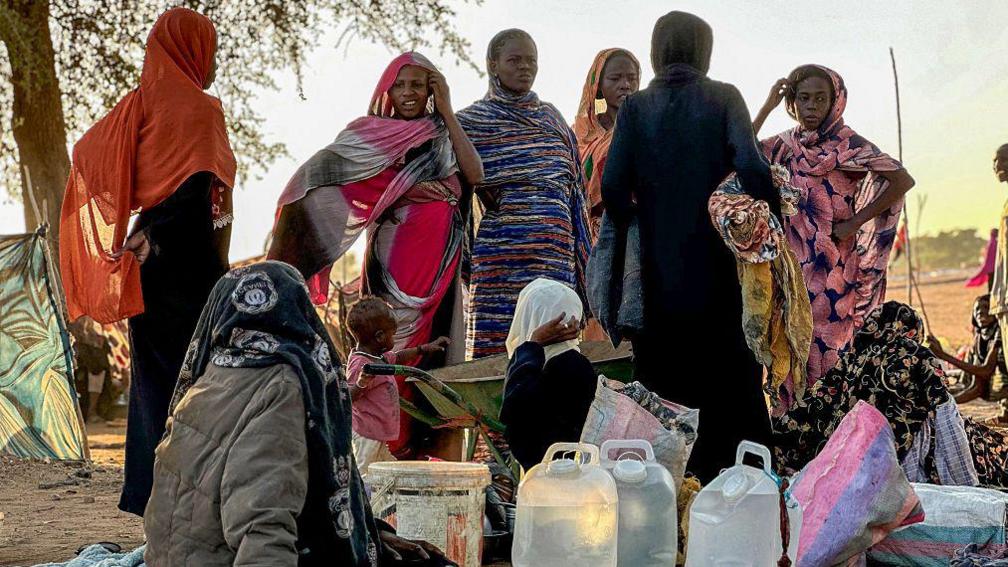 Displaced Sudanese who fled el-Fasher after the city fell to the Rapid Support Forces (RSF), rest near the the town of Tawila. Women in colourful scarves are standing up looking like they're wondering what to do next. Some people are sitting on the floor with their belongings.