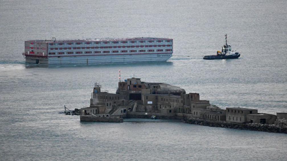 The Bibby Stockholm being towed by a tugboat. Part of the Dorset coast is in the foreground.