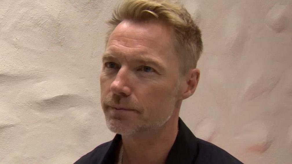 Ronan Keating wearing a black shirt and with short cropped blonde hair. He has a neutral expression his face and is pictured in front of a white wall. 