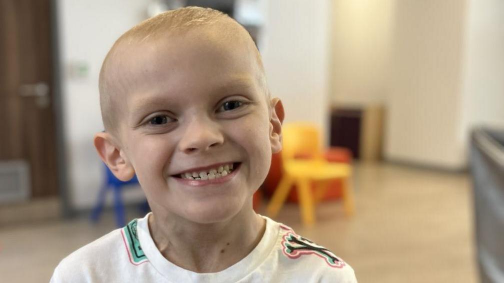 Coby is smiling at the camera in a waiting room at the hospital. He is wearing a white T-shirt with colourful graphics.