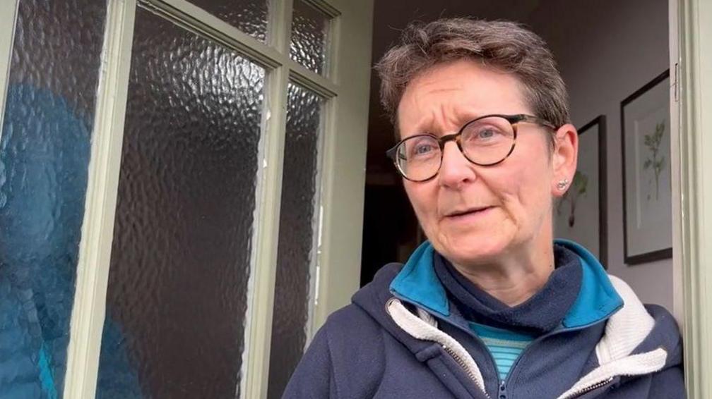 Julia Markham stands in the doorway of her home. She has short brown hair and is wearing a pair of glasses and a blue zip-up top.