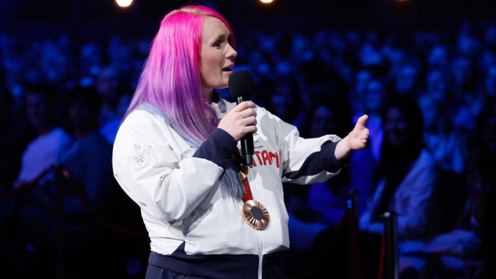 Jodie Grinham speaking with a microphone, wearing kit, at the event 