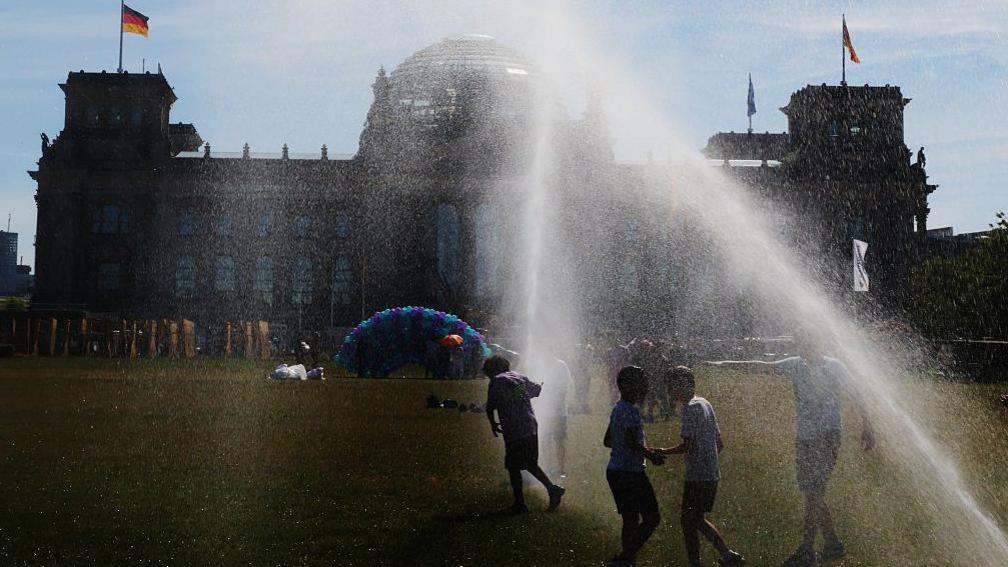 People pass by a watering system in front of the Reichstag on a hot summer day on June 24, 2026 