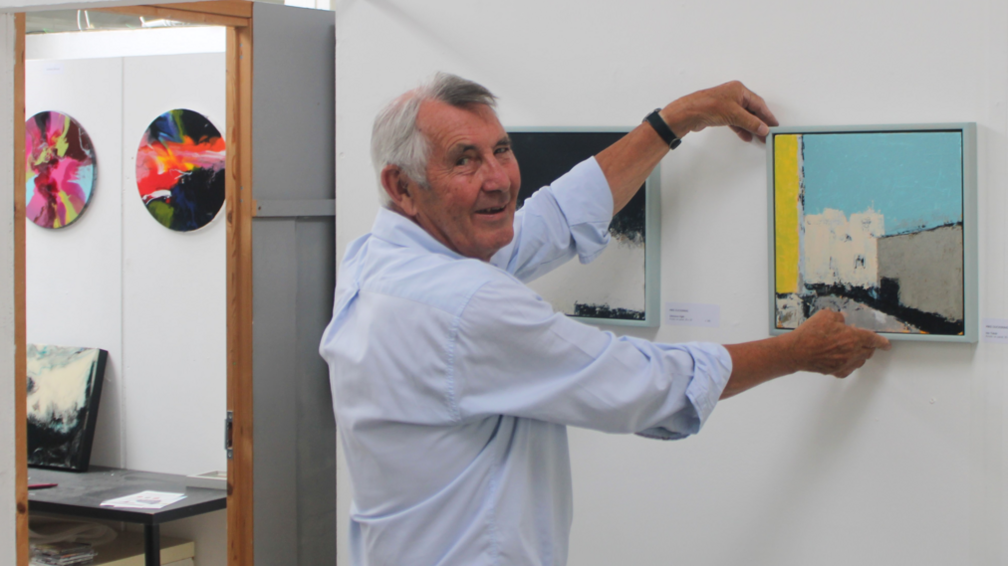 Mike, smiling to camera, holding up a small abstract painting in turquoise, grey and yellow