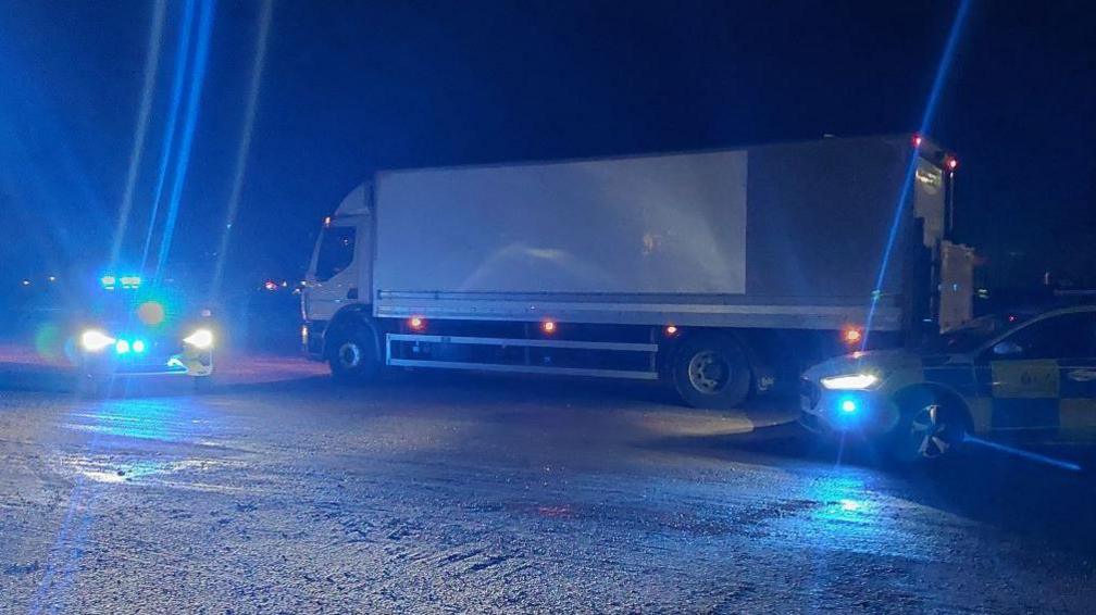 A white truck in the dark lit up by police car headlights. there are two police cars 