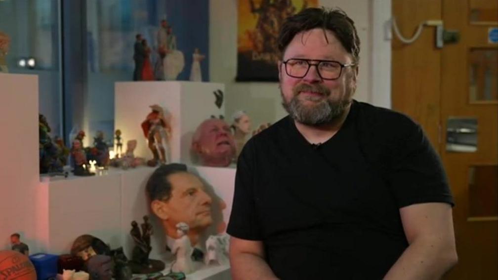 Tim Milward pictured sat down in a room surrounded by action figures. Tim is wearing a black crew neck t-shirt and thick framed black glasses/ He has dark brown hair that slightly flows over his ace and a full beard and moustache 