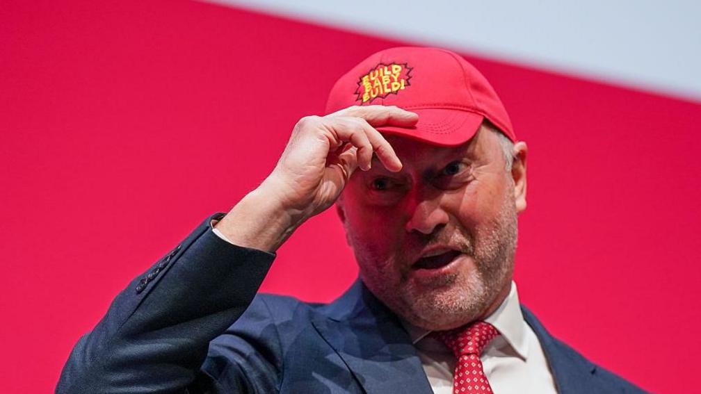 Steve Reed on the Labour party conference stage wearing a blue suit, red tie and red baseball cap with a yellow slogan on saying "build baby build".