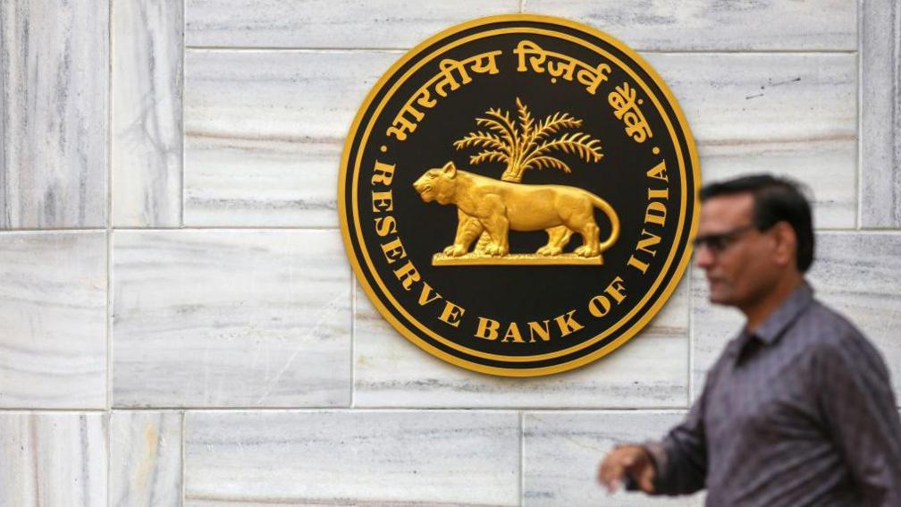 The image shows the logo for Reserve Bank of India (RBI) at the central bank's headquarters building in Mumbai with a spectacled man walking in the foreground.
