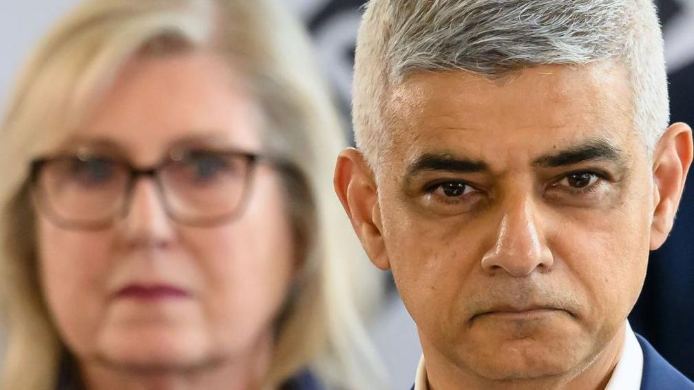 A close up of the Sir Sadiq Khan in the foreground, with Susan Hall out of focus behind him.
