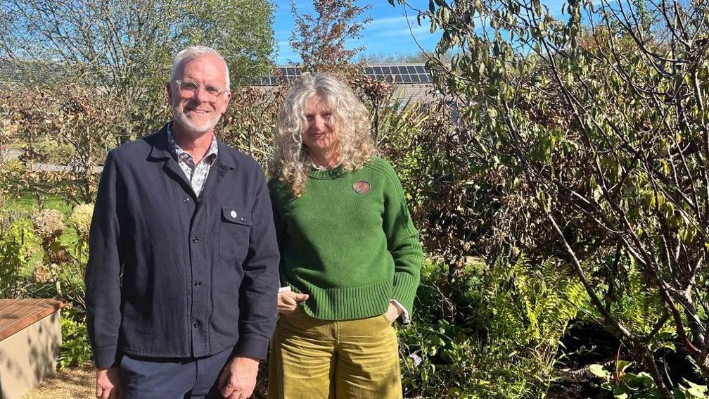 A man and a woman stand next to each other in a garden. The man is wearing a dark blue shirt and trousers, and the woman is wearing a dark green jumper with light green trousers. 