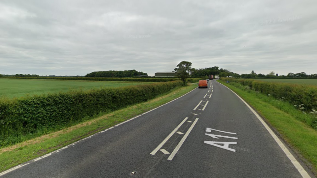 An A road in the middle of green fields. There is a red van driving away from the camera in the distance