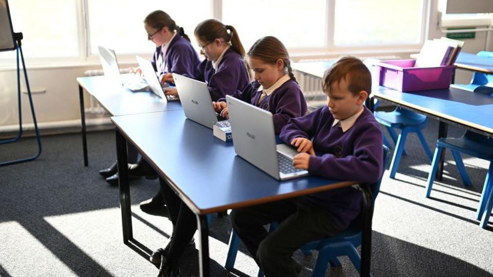 Four children in purple school uniforms do some school work on their laptops in a classrooms. 