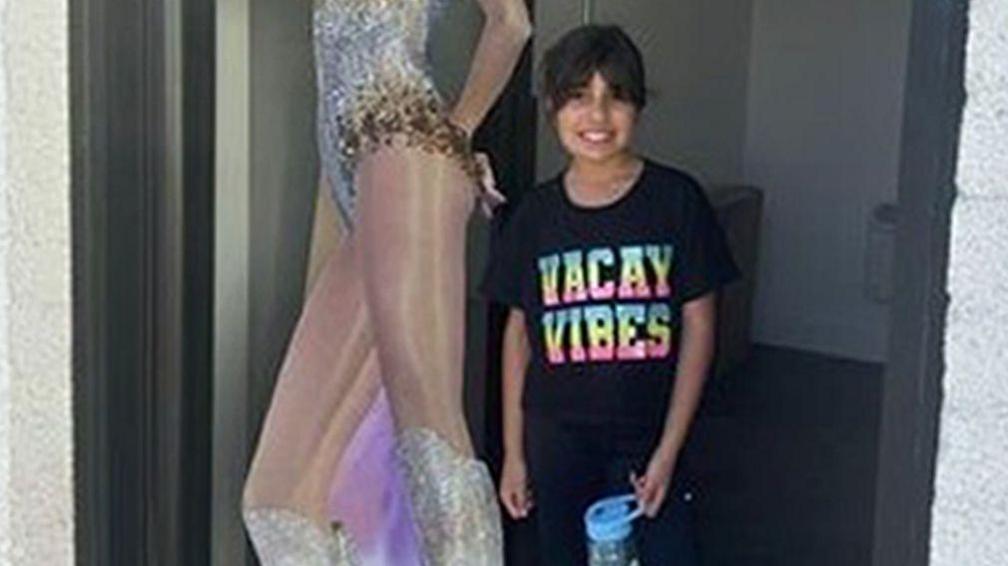 A smiling Alice stands outside the Hart Space dance studio next to a life-size cardboard cut-out of Taylor Swift, wearing a black t-shirt with the slogan 'Vacay Vibes' written in colourful letters. 