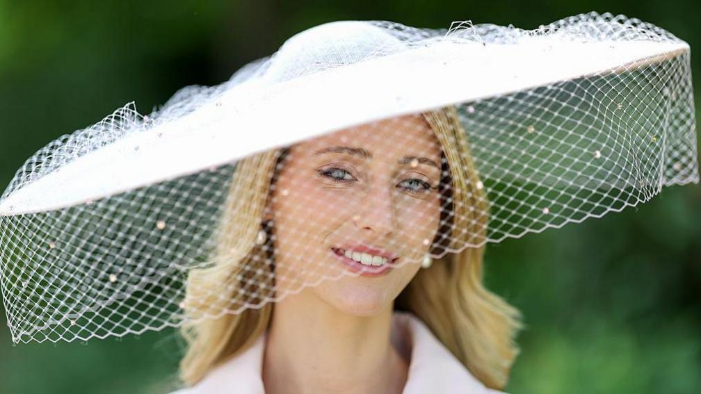 Flora Gibbs, who has blonde hair and pearl earrings, is pictured wearing a large hat with netting over the front of it.