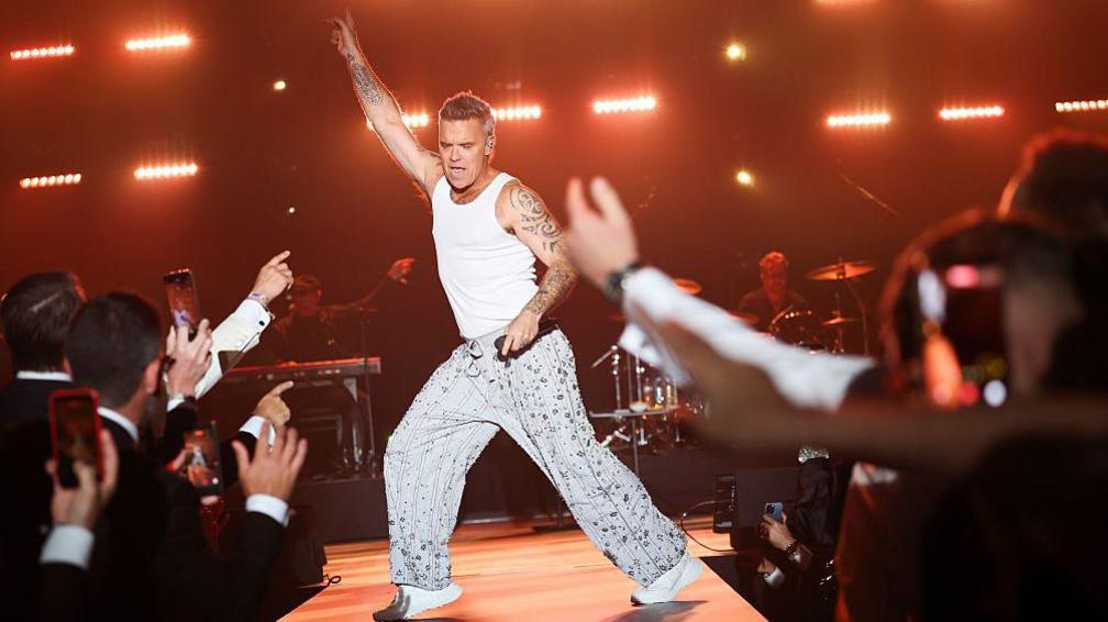 Robbie Willians dancing on stage and holding a microphone. People in the crowd are cheering and taking photos.